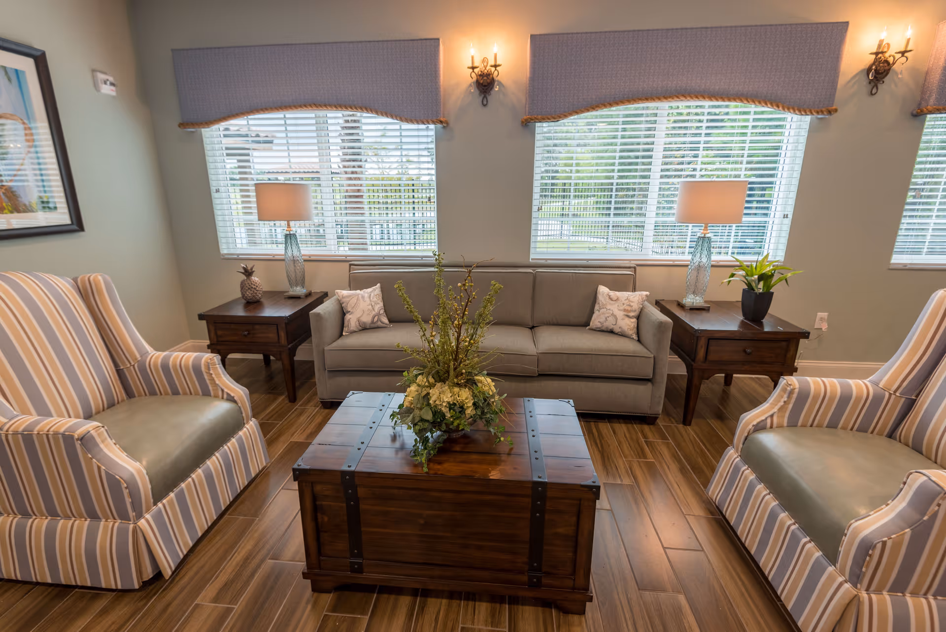 A cozy living room with a gray sofa flanked by two wooden side tables, each holding a lamp and decorative items. Two striped armchairs face the sofa, and a wooden coffee table with a floral arrangement sits in the center. Large windows with blinds and valances allow natural light to fill the room.