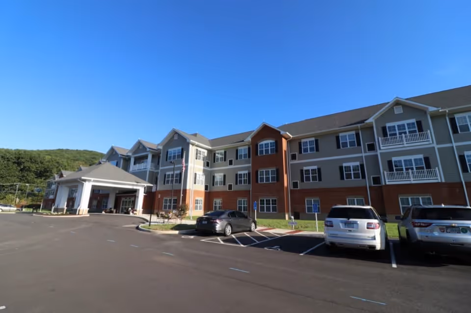 Three-story senior living facility with a covered entrance, adjacent parking lot and cars under a clear blue sky.