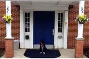 Brick building entrance with blue double doors flanked by white columns and hanging flower baskets, and a black dog sitting on a dark mat.