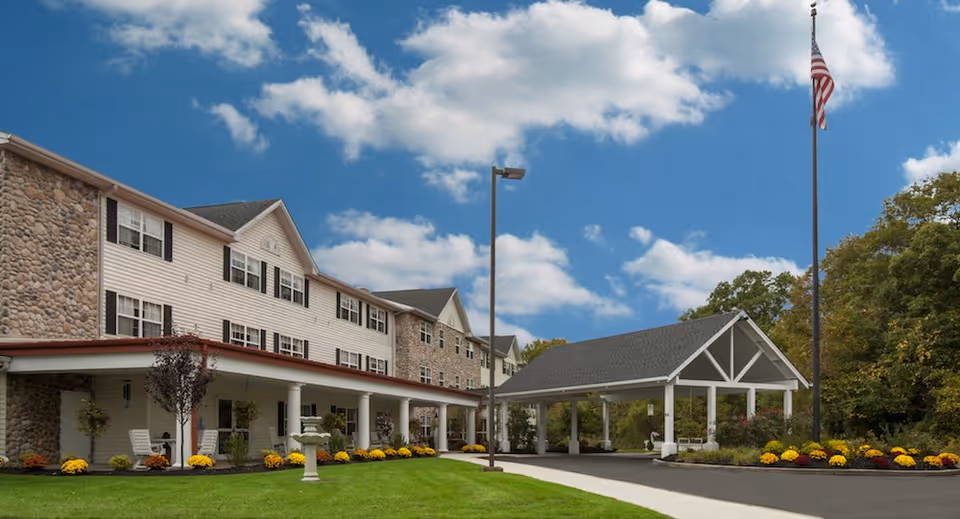 Exterior view of a senior living facility building with stone and white siding, a covered entrance with white pillars, a flagpole with an American flag, and a well-maintained lawn with yellow flowers under a partly cloudy blue sky.