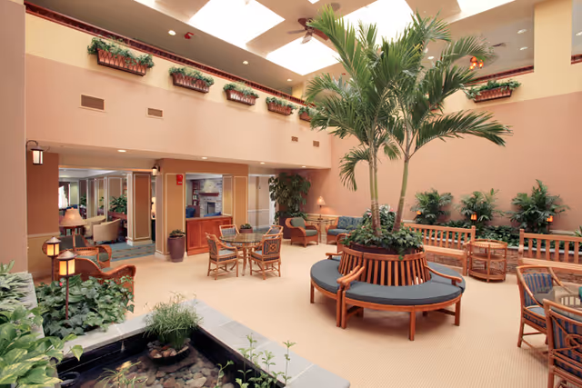 Sunlit indoor atrium lounge with a central round planter bench, palm trees, wicker chairs and tables, and a small water feature.