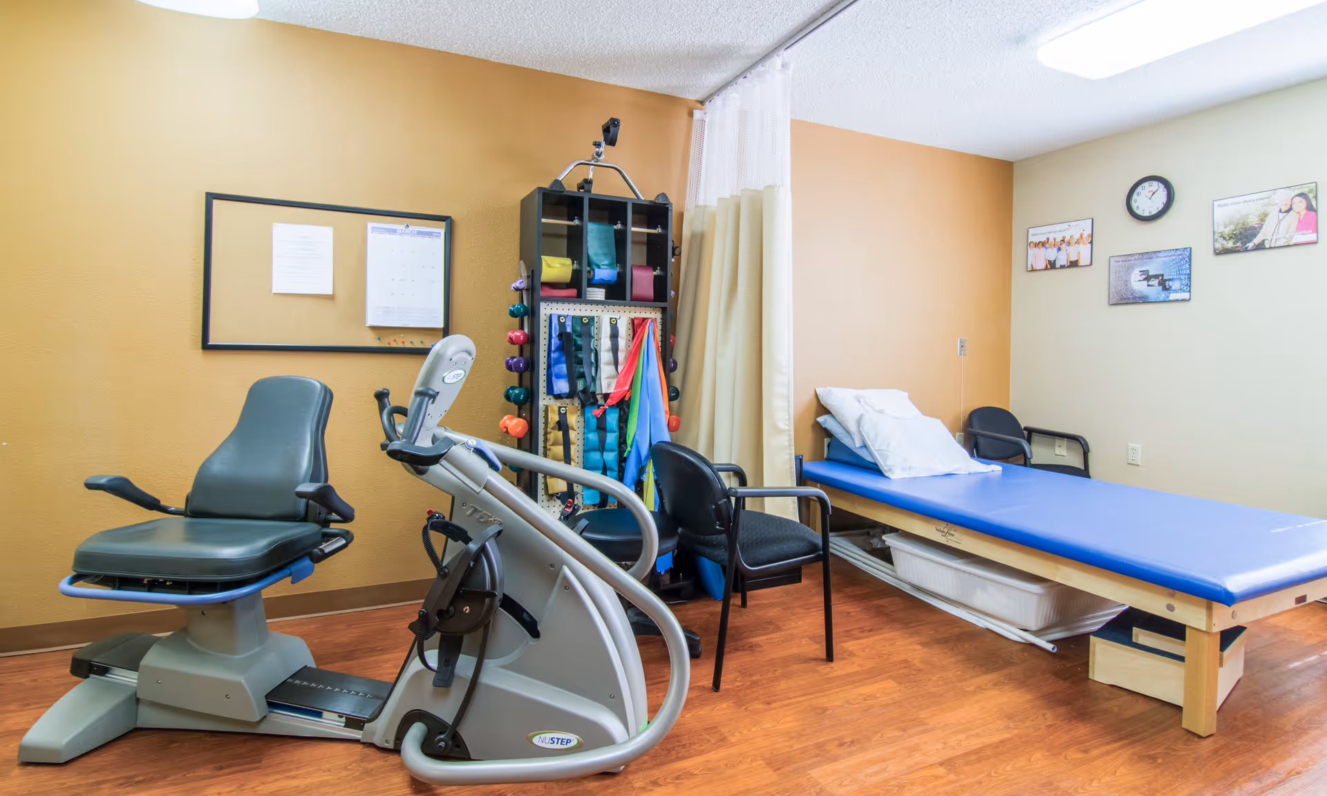 A physical therapy room with a recumbent exercise bike, a treatment table with blue padding and white pillows, two black chairs, a rack holding colorful resistance bands and small weights, a corkboard with papers, and a wall clock above framed pictures.