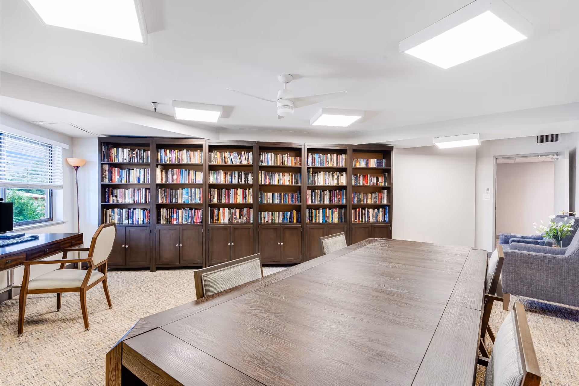 Common room with a long wooden table and chairs facing built-in bookshelves, a desk by the window, and a small seating area.