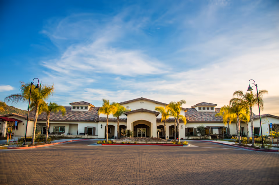 Front exterior view of Manzanita Village facility with a wide driveway, palm trees, and a building with a tiled roof under a partly cloudy sky.