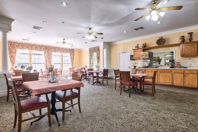 A bright dining room with multiple wooden tables and chairs arranged neatly. The tables are covered with pink tablecloths and have small flower vases as centerpieces. Large windows with floral curtains allow natural light to fill the room. Ceiling fans with lights hang from the ceiling. There is a serving area with wooden cabinets and a pass-through window to the kitchen.