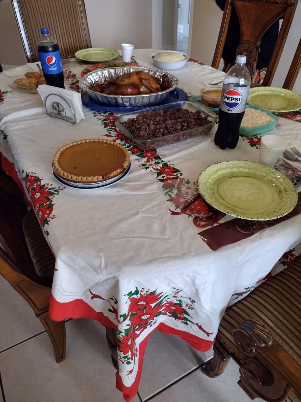 A dining table set for a meal with a white tablecloth decorated with red poinsettias. On the table are plates, a roasted chicken in a foil tray, a glass dish with ground meat, a pumpkin pie, two bottles of Pepsi, a bowl of mashed potatoes, and some other side dishes. There are wooden chairs around the table.