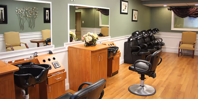 Interior of a salon area in an assisted living facility featuring multiple black salon chairs in front of wooden counters with sinks, large mirrors on the wall, and a couple of beige armchairs in the background. The walls are painted green with white wainscoting and decorated with framed artwork and a floral wall sculpture.