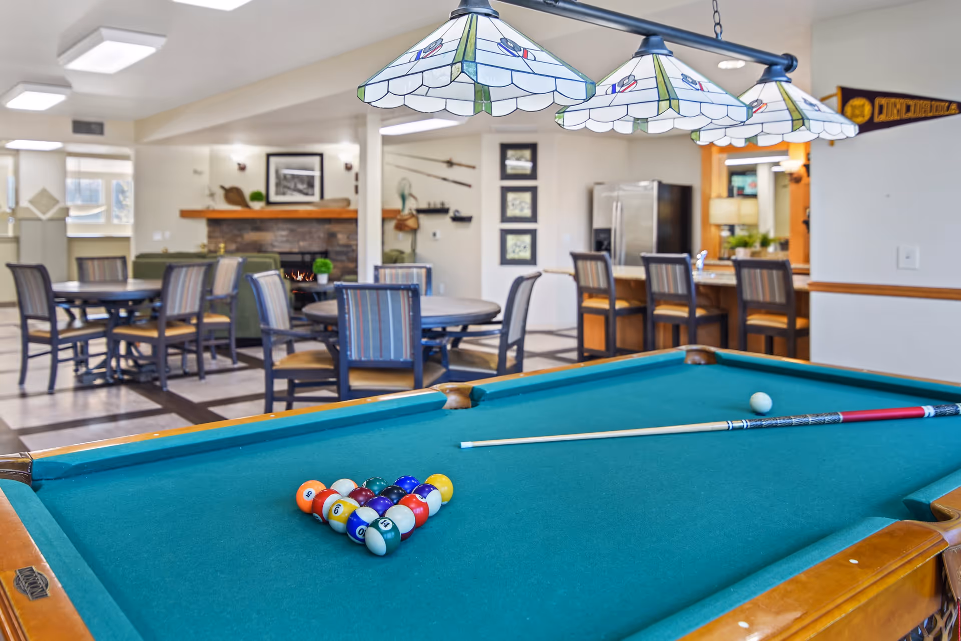A billiards table with a triangle of colored balls and a cue stick in a recreational room featuring dining tables and a fireplace in the background.