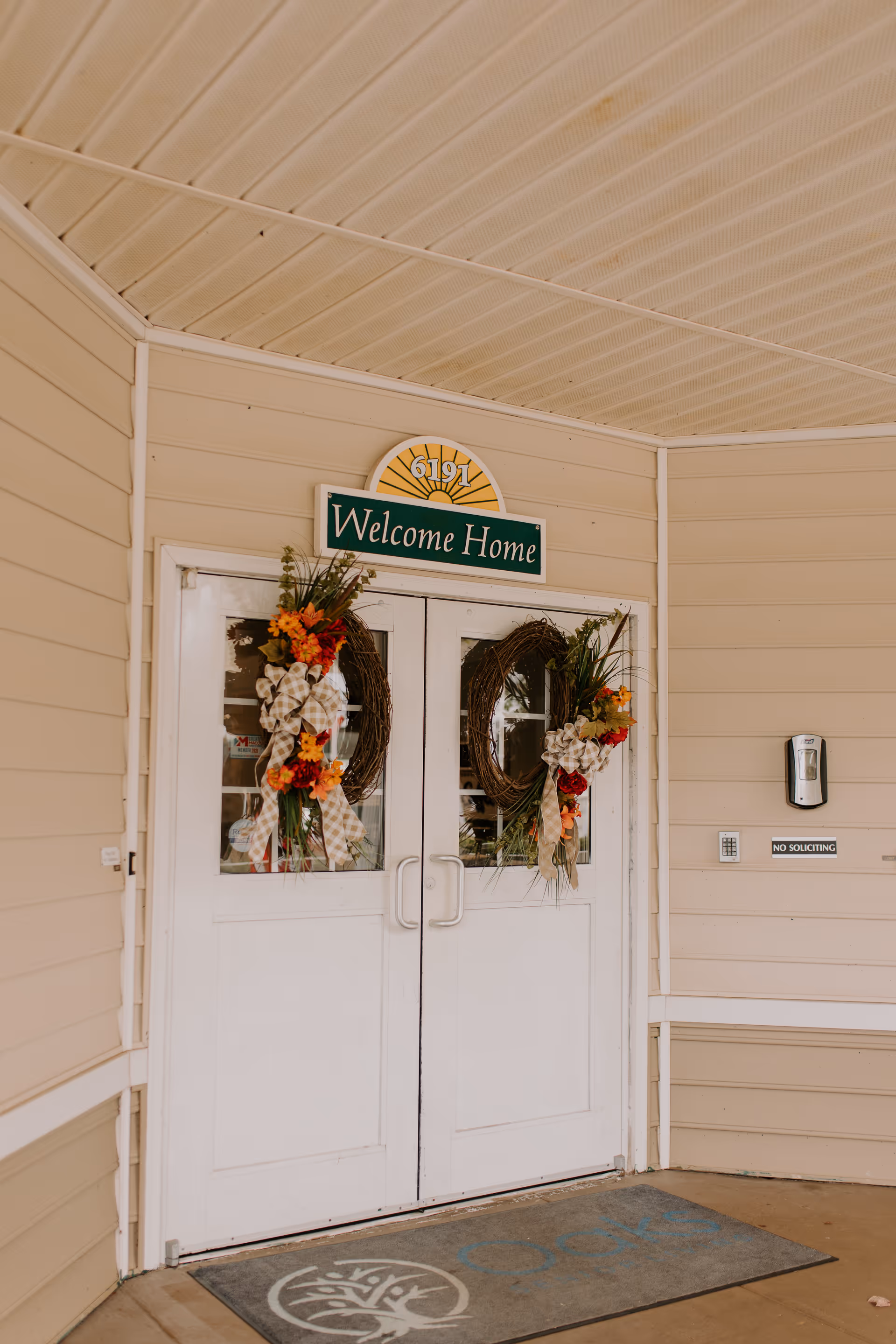 Double white doors decorated with floral wreaths and a sign above reading 'Welcome Home' with the number 6191. The entrance has beige siding walls, a keypad entry system, a hand sanitizer dispenser, and a doormat with the Oaks at Lake Wildwood logo.
