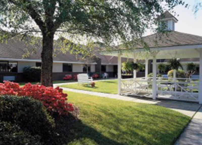 Outdoor courtyard area with a large tree, green grass, red flowering bushes, a white gazebo with hanging plants, and a building with windows in the background under a clear sky.