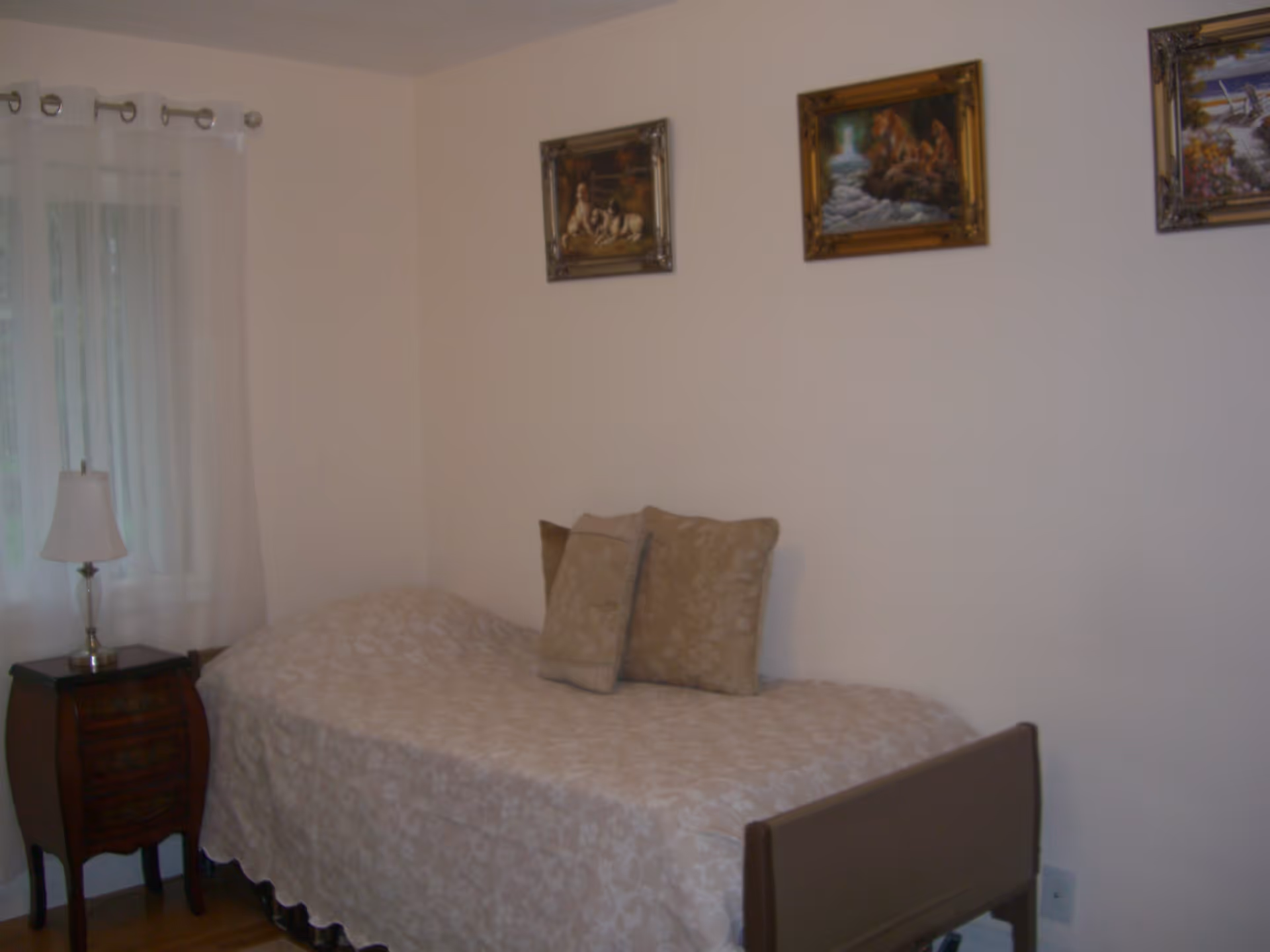 A small bedroom with a single bed covered in a beige floral bedspread and two matching pillows. Next to the bed is a wooden nightstand with a white lamp. The wall above the bed has three framed paintings. A window with sheer white curtains is visible on the left side.