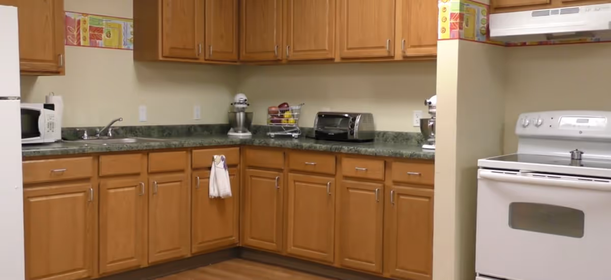 A kitchen with wooden cabinets, green countertops, a white microwave, a toaster oven, a stand mixer, a white stove with an oven, and a sink with a faucet. There are some kitchen towels hanging on a cabinet handle and a fruit basket on the counter.