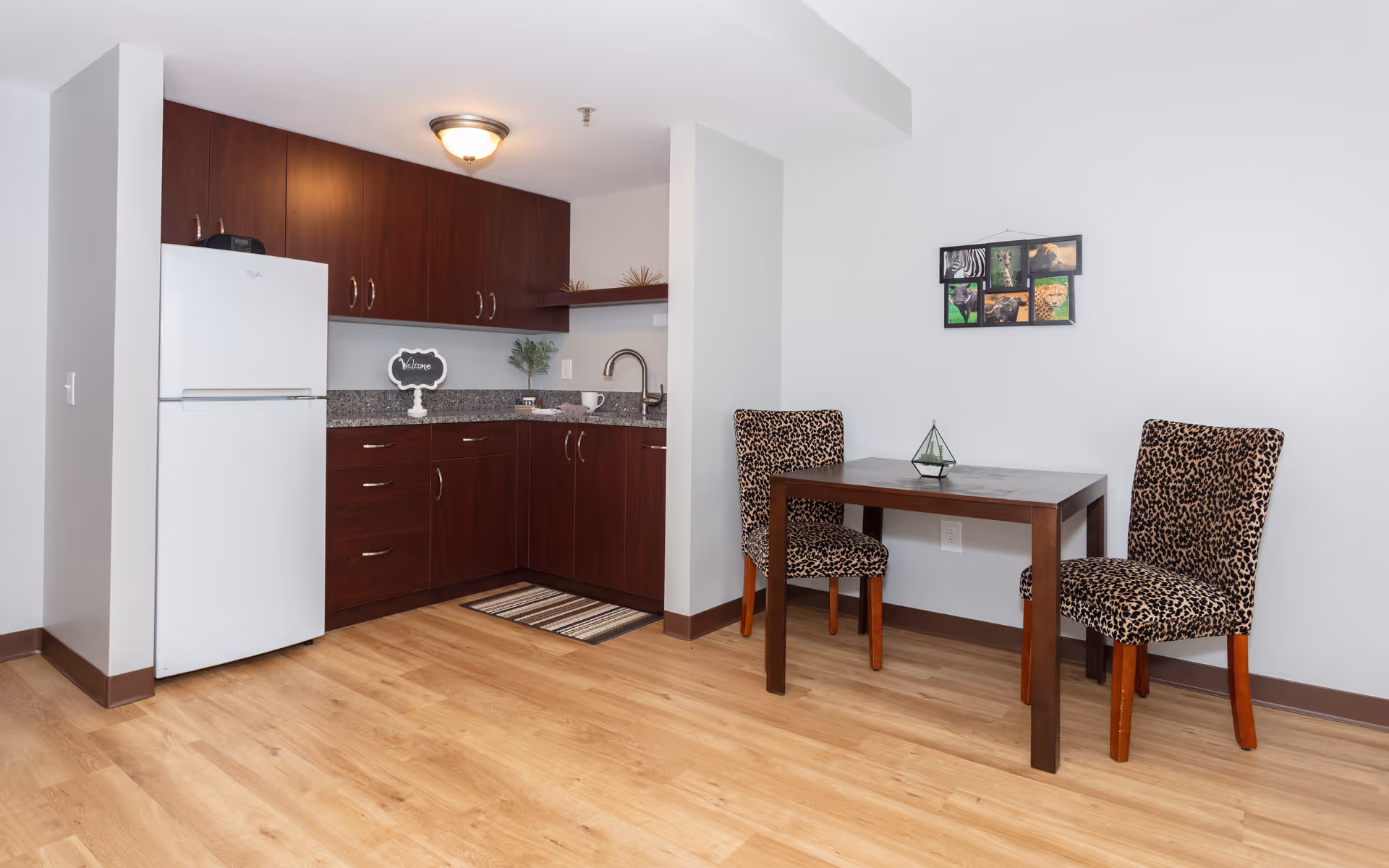 A small kitchen area with dark wooden cabinets, a white refrigerator, granite countertops, and a stainless steel sink. Adjacent to the kitchen is a wooden dining table with two leopard-print upholstered chairs. A small framed collage of animal photos hangs on the white wall above the table. The floor is light wood, and the space is well-lit with a ceiling light fixture.