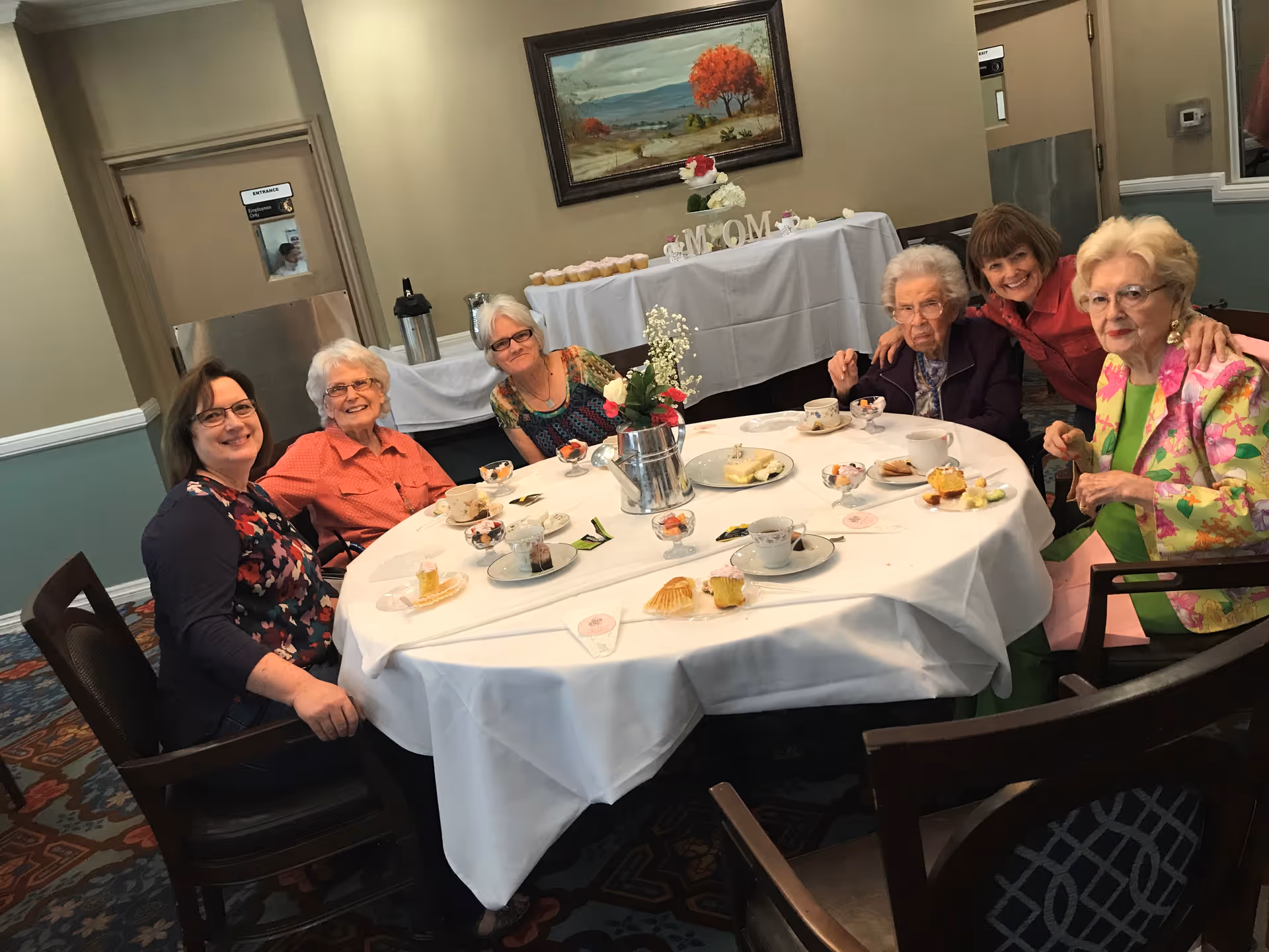 Six elderly women sitting around a round table covered with a white tablecloth, enjoying tea and desserts in a dining room. There is a flower centerpiece on the table and a buffet table with cupcakes and a 'MOM' decoration in the background. The room has beige walls and a painting of a landscape with a red tree.