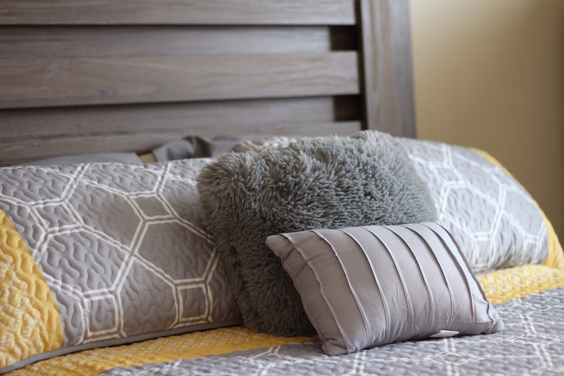 Close-up view of a bed with a wooden headboard, featuring a yellow and gray patterned quilt and two decorative pillows, one fluffy gray and one smooth gray with pleats.