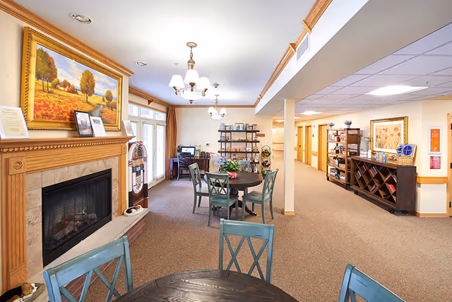 A cozy common area in a senior living facility featuring a fireplace with a large framed painting above it, round tables with chairs, a jukebox, shelves with books and decorative items, and a hallway leading to other rooms. The space is warmly lit with ceiling lights and has carpeted floors.