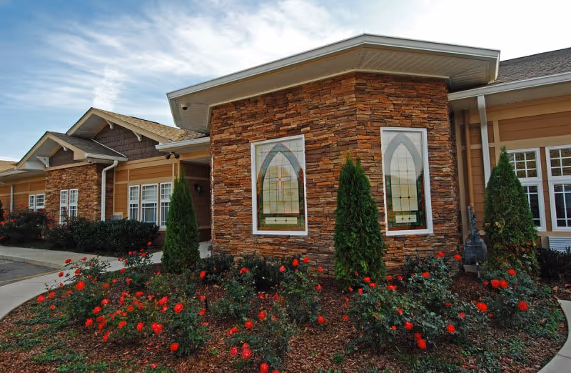 Front exterior of a single-story building with a stone facade, arched stained-glass windows and a landscaped bed of red roses and shrubs.
