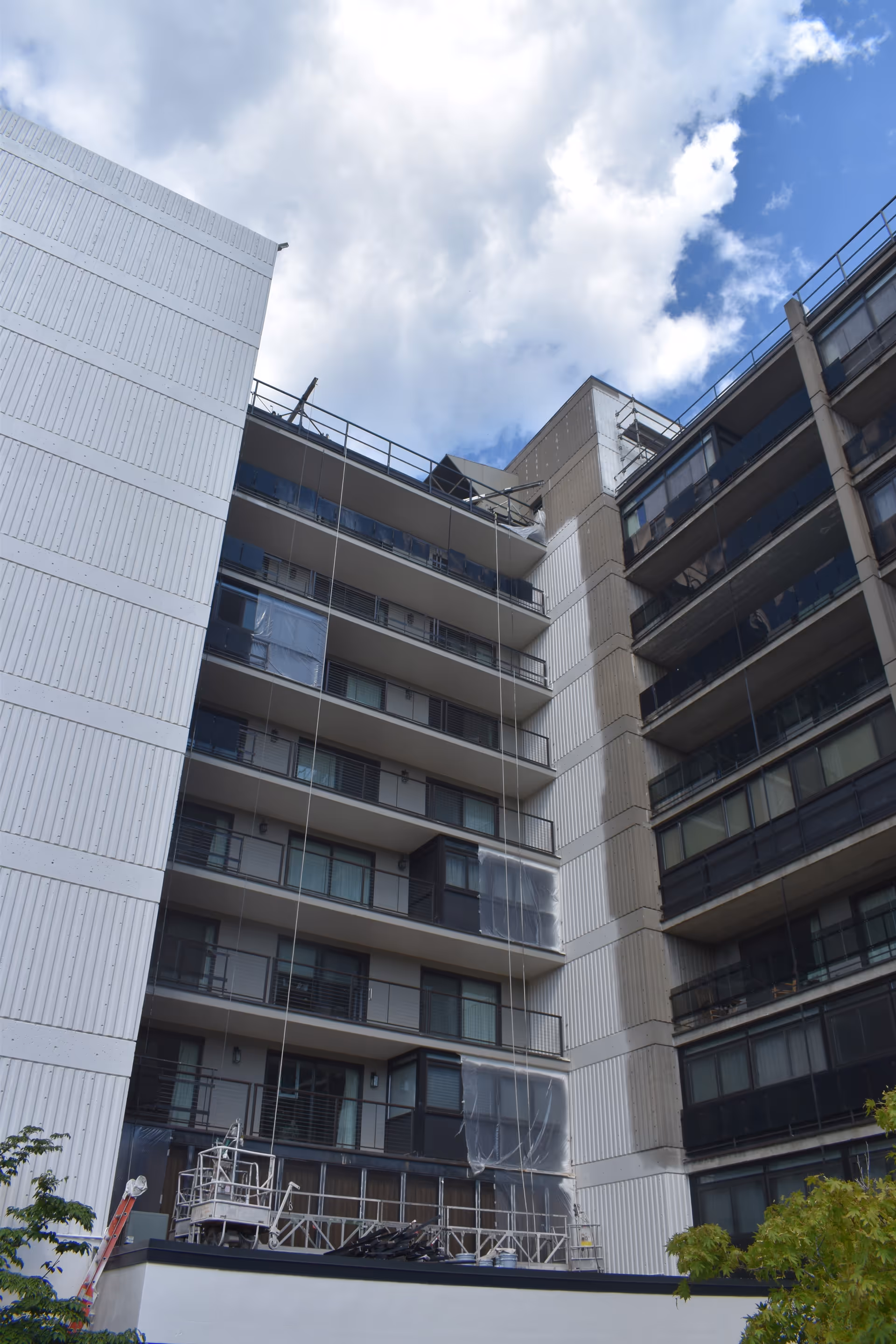Exterior view of a multi-story assisted living building with balconies, scaffolding and protective sheeting under a partly cloudy sky.