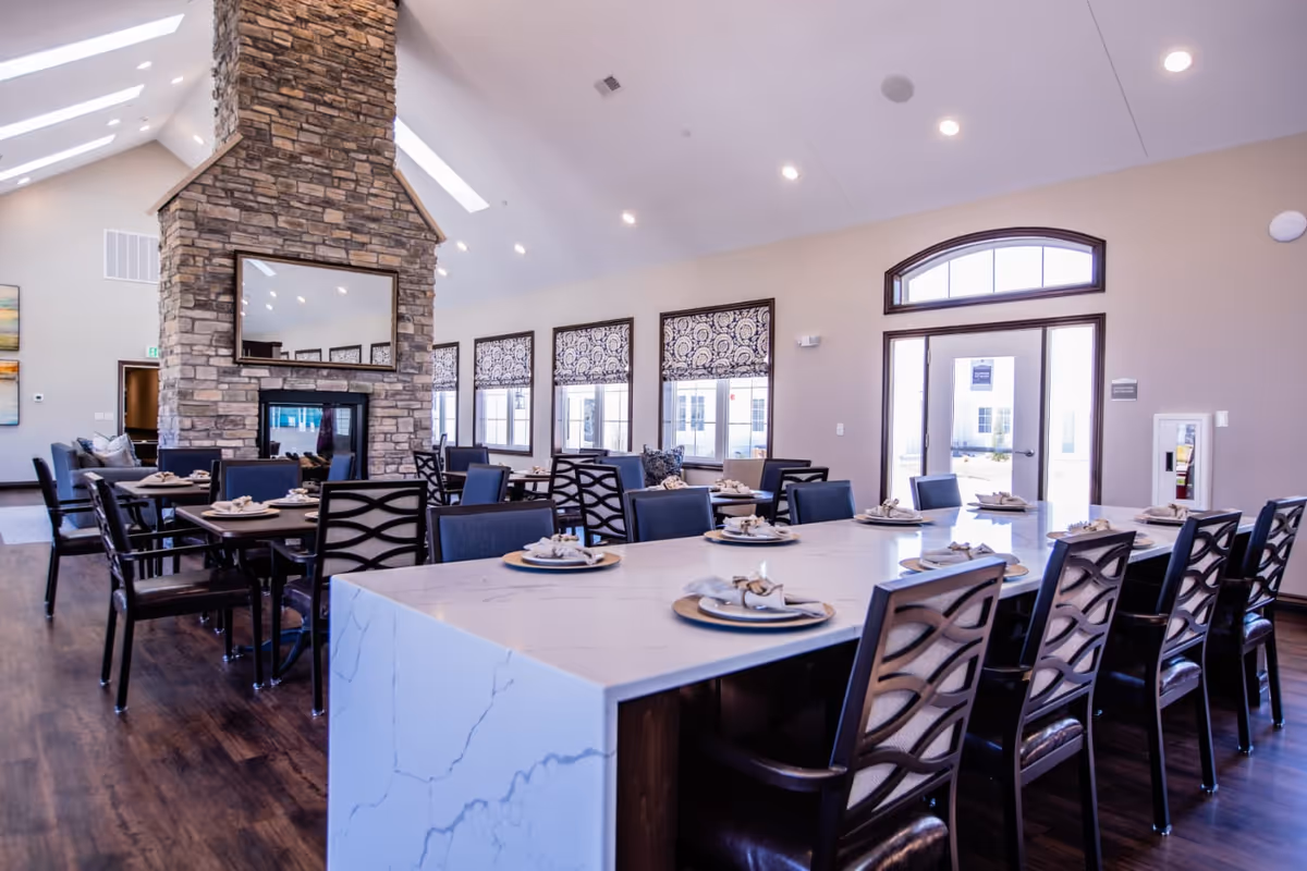 Spacious dining room with a long marble-topped communal table set for meals, multiple chairs, a stone fireplace, and large windows.