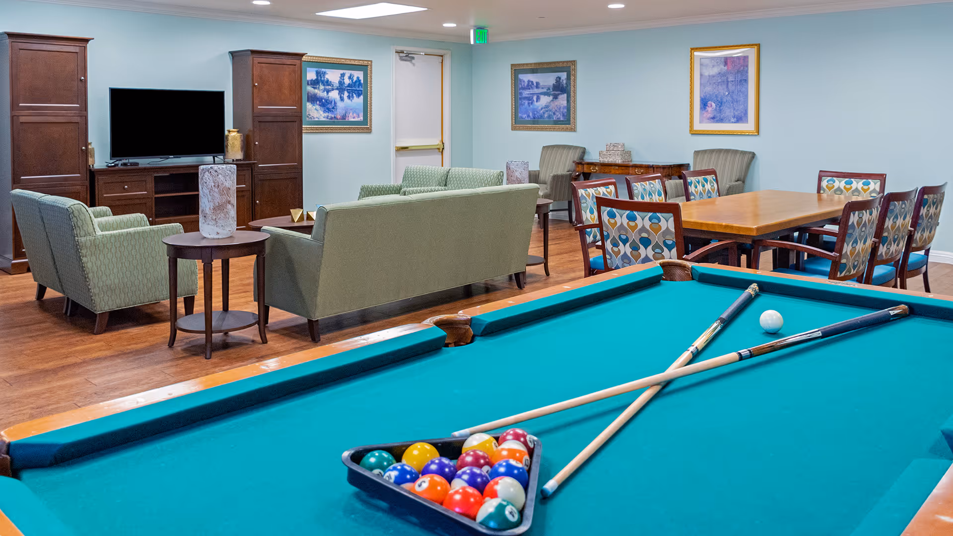 A recreational room with a pool table in the foreground, two pool cues crossed on the table, and a rack of colorful billiard balls. In the background, there is a seating area with green upholstered sofas and armchairs, a wooden TV cabinet with a flat-screen television, a wooden dining table with patterned chairs, and framed artwork on light blue walls.