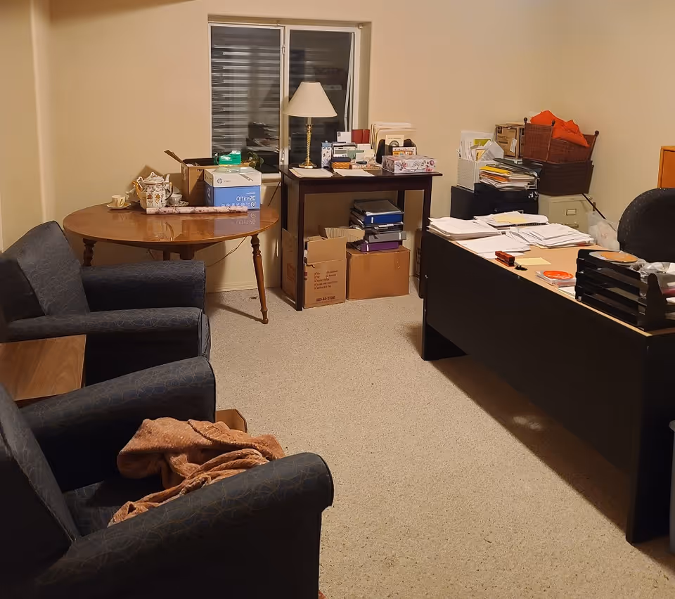Cluttered interior room with upholstered armchairs, a round table and a desk covered in papers, boxes and a lamp beneath a window.