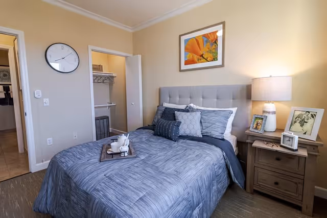 A cozy bedroom with a neatly made bed featuring gray bedding and multiple pillows. A wooden nightstand with a lamp and framed photos sits beside the bed. A colorful floral painting hangs on the beige wall above the headboard. An open closet and a doorway leading to a bathroom are visible in the background.