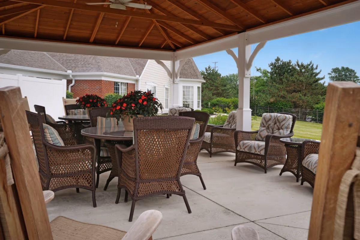 Covered outdoor patio area with wicker chairs and tables, some with cushions and potted red flowers on the tables, overlooking a grassy yard with trees and a white fence.
