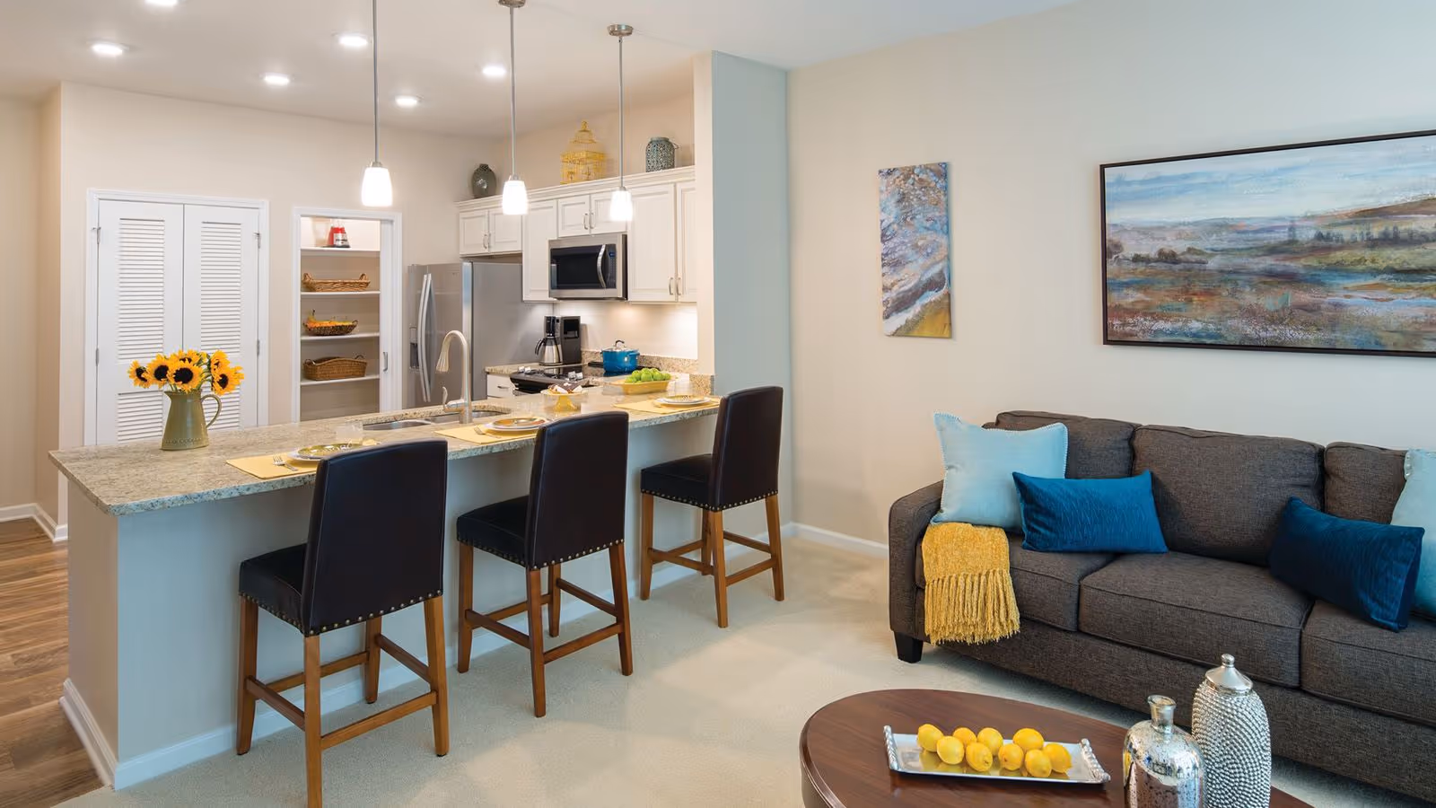 A modern open-concept kitchen and living room area in a senior living community. The kitchen features a granite countertop island with three dark leather bar stools, pendant lighting, stainless steel appliances including a refrigerator and microwave, and an open pantry with shelves. The living room has a dark gray sofa adorned with blue and light blue pillows and a yellow throw blanket. A round wooden coffee table holds decorative items and a tray with lemons. Two abstract paintings hang on the wall above the sofa.