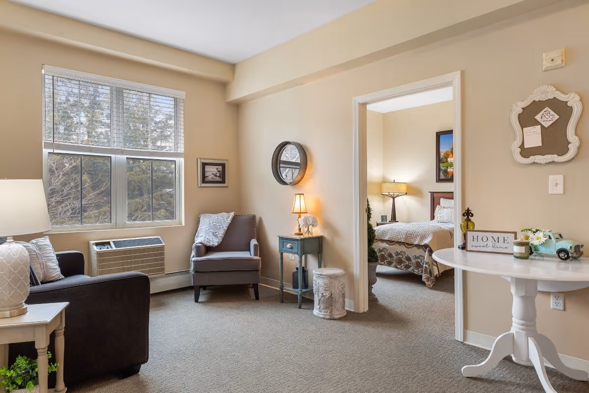 A cozy living room area with a dark armchair, a gray armchair with a throw blanket, a small side table with a lamp, and a window with blinds showing trees outside. Adjacent to the living room is a bedroom with a bed, nightstand, and lamp. A white round table with decorative items and a 'Home sweet home' sign is positioned near the entrance to the bedroom.