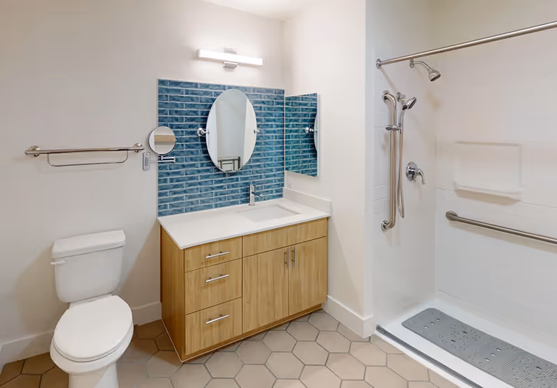 Modern accessible bathroom with a wooden vanity, blue tile backsplash and round mirror, toilet, and a roll-in shower with grab bars.