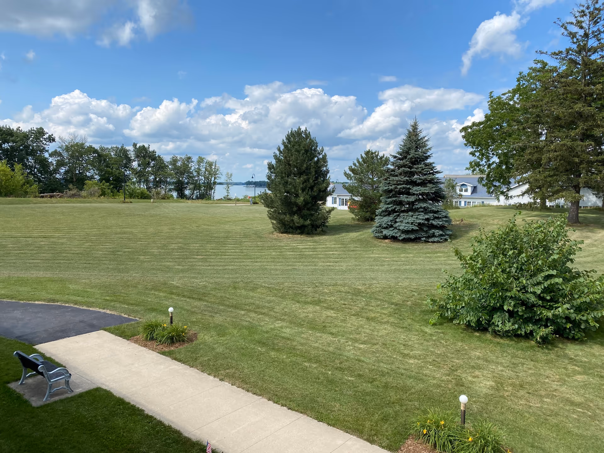 A large, well-maintained grassy lawn with several trees and bushes under a partly cloudy blue sky. A concrete walkway runs through the lawn with a bench beside it. In the background, there is a glimpse of a body of water and a white building with a gray roof partially visible behind the trees.