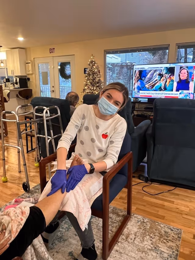 A caregiver wearing a face mask and purple gloves is seated in a chair, gently holding and massaging the foot of an elderly person who is covered with a blanket. The setting is a cozy living room with wooden floors, a Christmas tree decorated with lights, a television showing a news program, and mobility aids like walkers visible in the background.