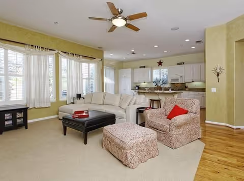 A spacious living room with a beige sectional sofa, a patterned armchair with a red cushion, and a matching ottoman. There is a black coffee table with a red book on it. The room has large windows with white curtains, a ceiling fan with lights, and an open view into a kitchen area with white cabinets and a breakfast bar with stools.