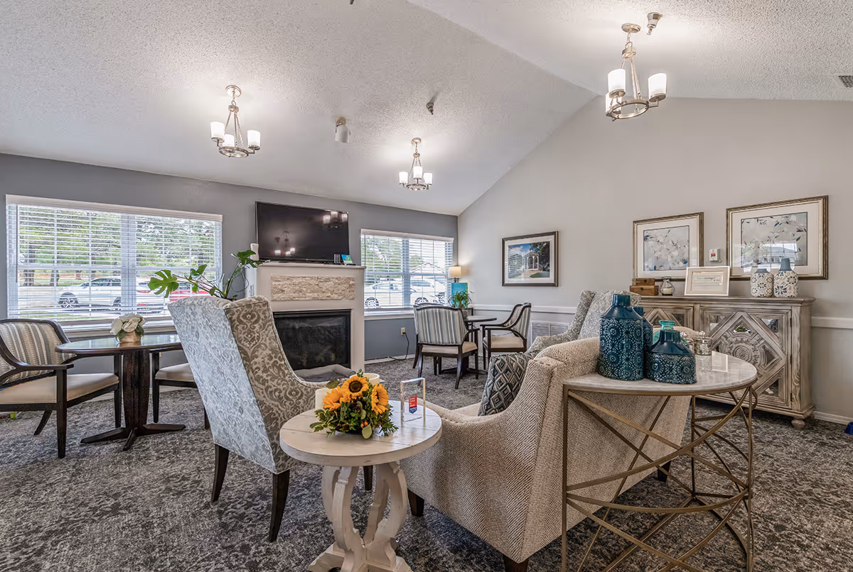 A well-lit communal living room with upholstered armchairs, tables, a fireplace with TV above, and decorative accents.