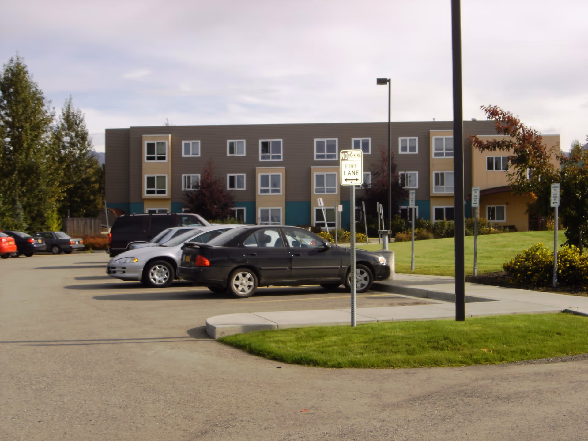 Parking lot with several parked cars in front of a three-story building with multiple windows, surrounded by grass, trees, and a few signs including a 'No Parking Fire Lane' sign.