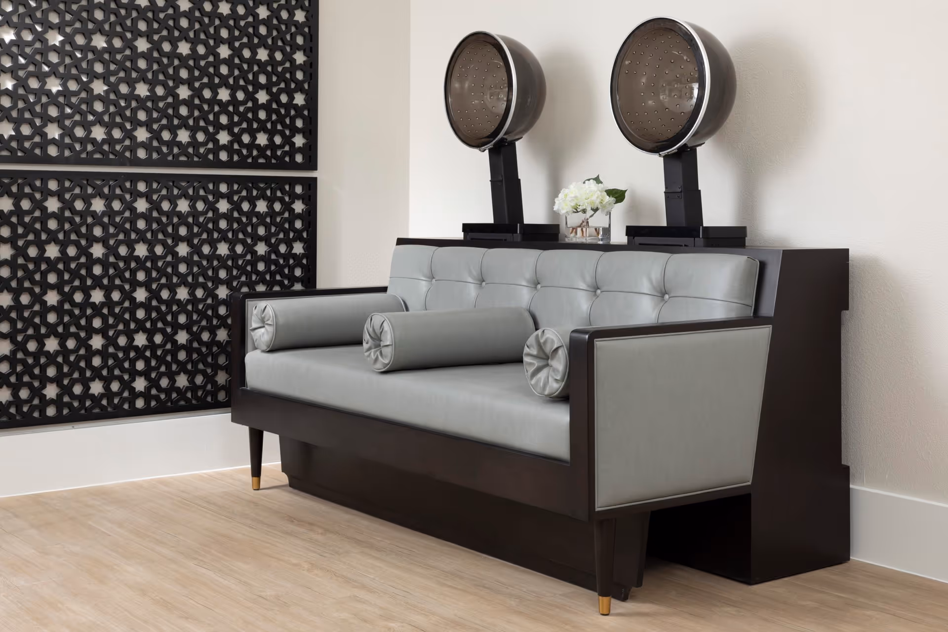 A modern salon area with a gray leather tufted sofa featuring three cylindrical bolster pillows. Behind the sofa are two vintage-style hair dryers mounted on a dark wooden console table, with a small vase of white flowers placed between them. The wall to the left has decorative black lattice panels, and the floor is light wood.