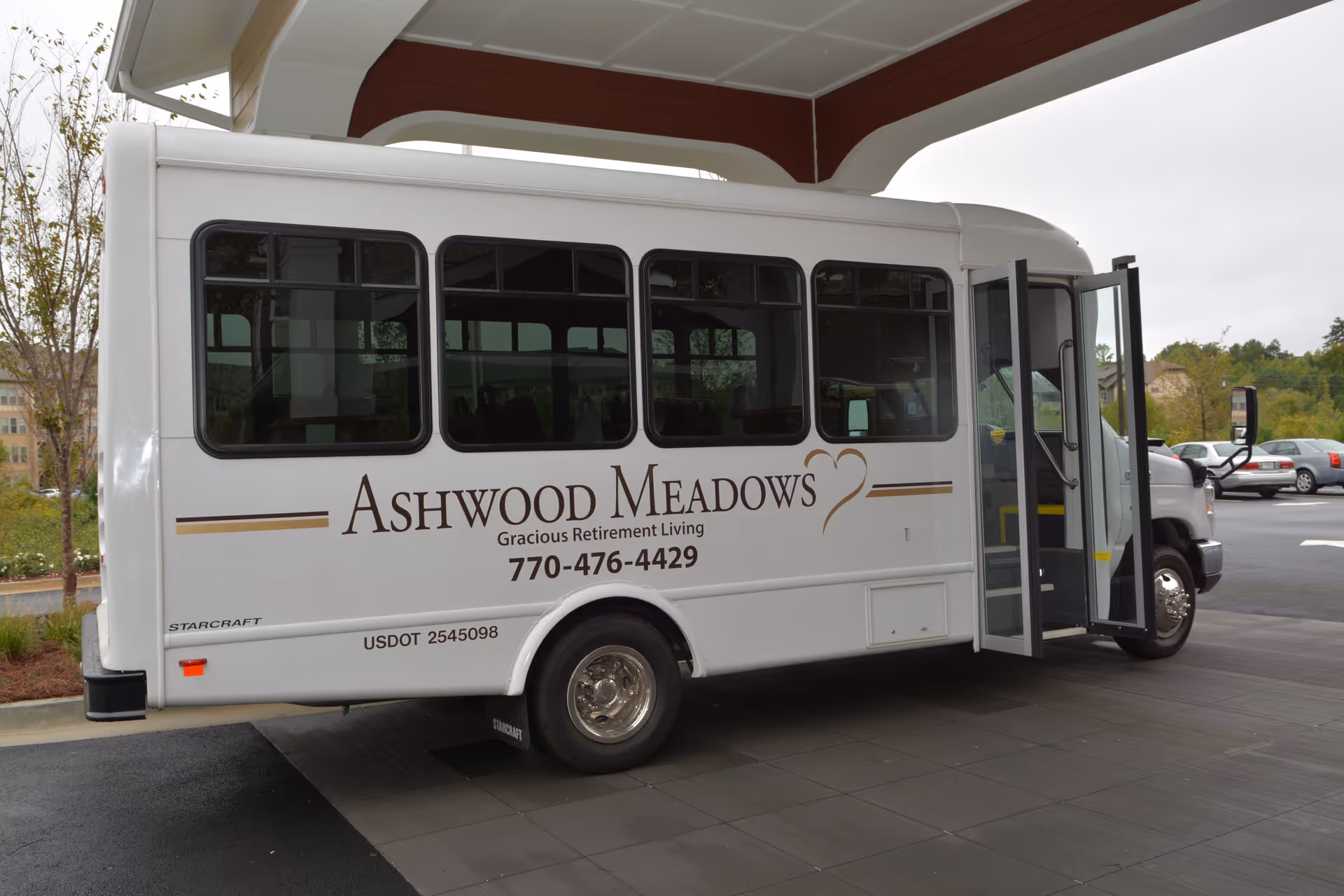 A white Ashwood Meadows retirement community shuttle bus parked under a covered entrance with its door open.