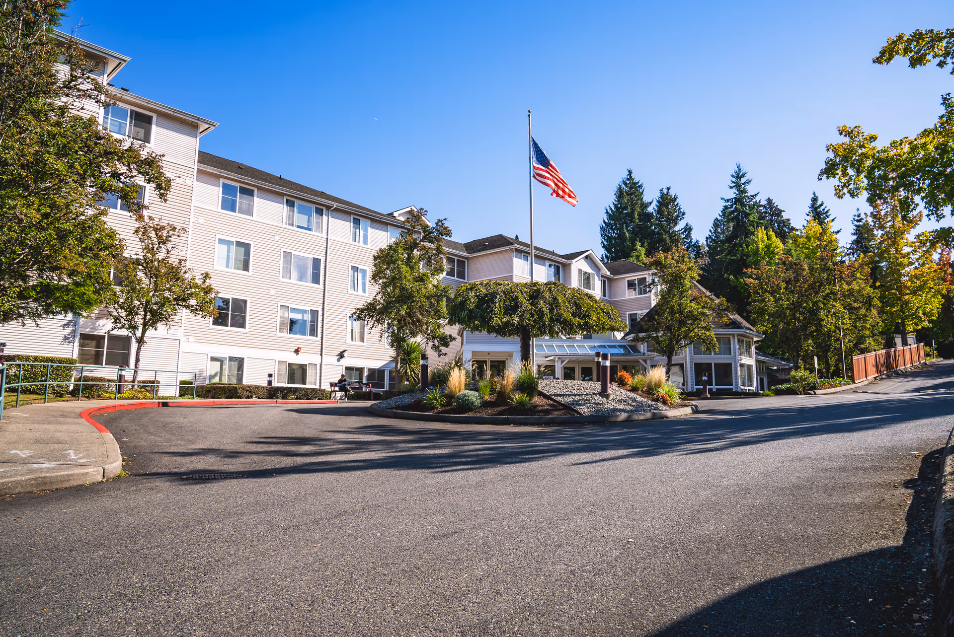 Exterior view of a multi-story assisted living facility with beige siding, surrounded by trees and landscaping. An American flag flies on a flagpole in front of the building, with a paved driveway and a roundabout in the foreground under a clear blue sky.