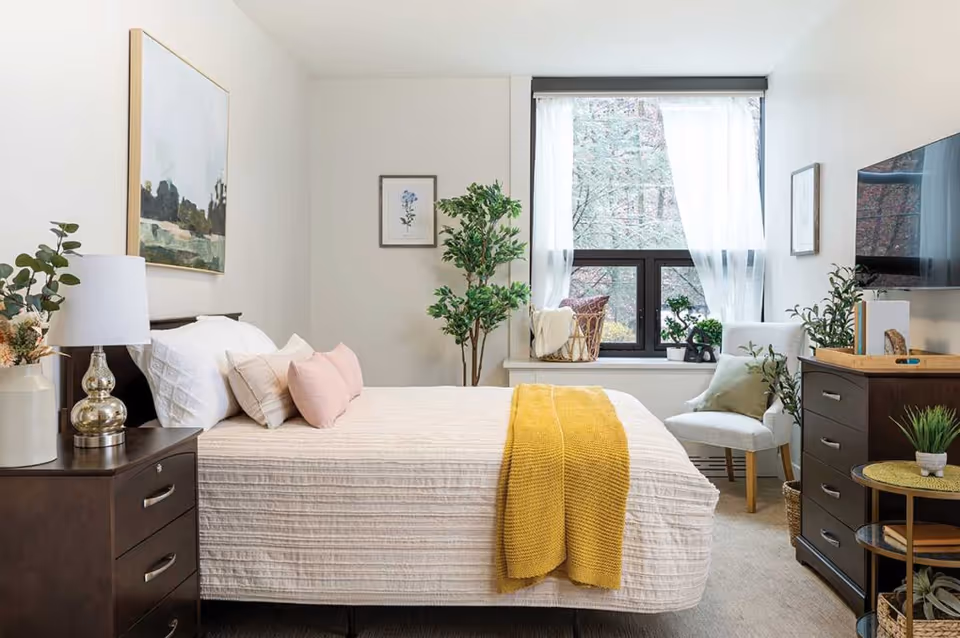 A bright and cozy bedroom with a neatly made bed featuring white and pink pillows and a yellow throw blanket. There is a dark wooden nightstand with a lamp and a vase with flowers on the left side of the bed. A large window with sheer white curtains lets in natural light, and there is a white armchair with a green pillow next to a dark wooden dresser with a TV mounted above it. The room is decorated with framed artwork and green plants.