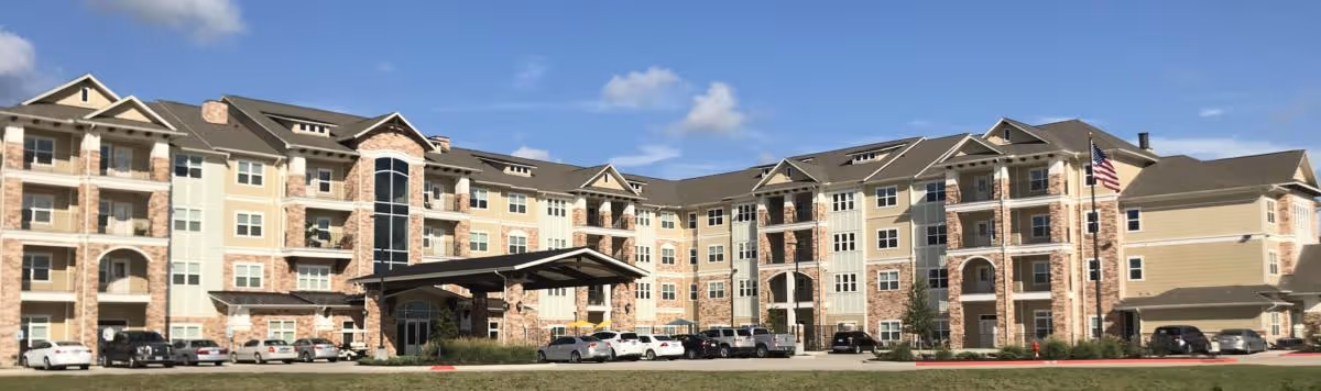 Exterior view of a large multi-story senior living facility building with a covered entrance, multiple windows, balconies, and an American flag on a flagpole. Several cars are parked in front of the building under a clear blue sky with some clouds.