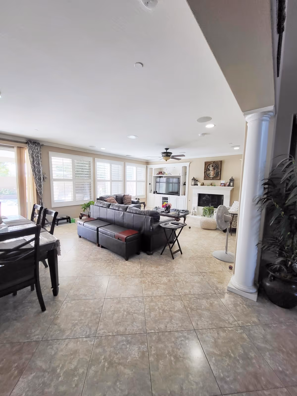 A spacious living room with large windows covered by white shutters allowing natural light to fill the room. The room features a dark leather sectional sofa with cushions, a coffee table, a side table with a remote control, and a standing fan. There is a fireplace with decorative items on the mantel and a built-in entertainment center with a television. The floor is tiled, and there is a dining table with chairs visible on the left side. A white column and a potted plant are on the right side of the image.