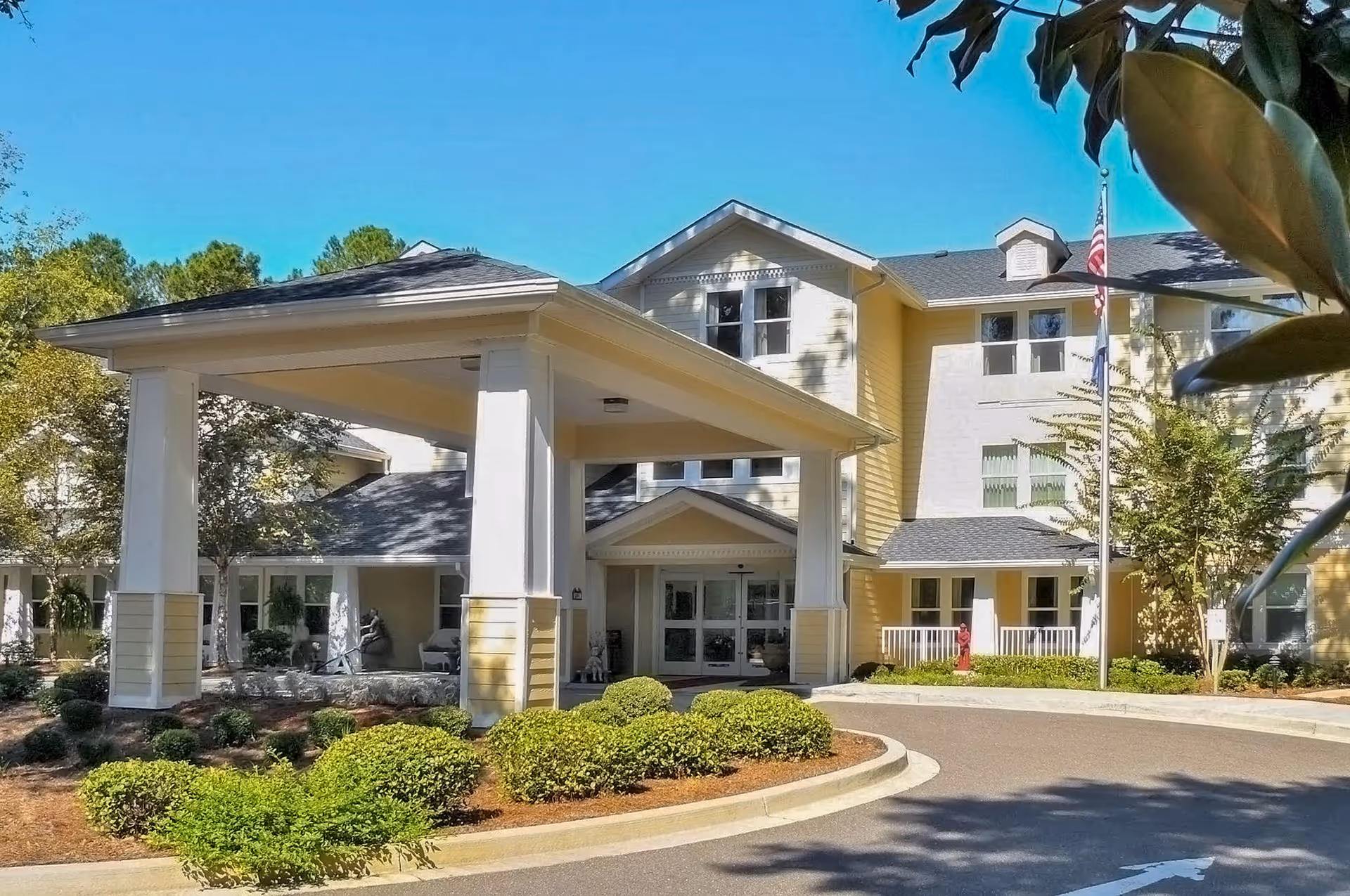 Yellow multi-story senior living facility with a covered entrance, flagpole, and neatly trimmed landscaping.
