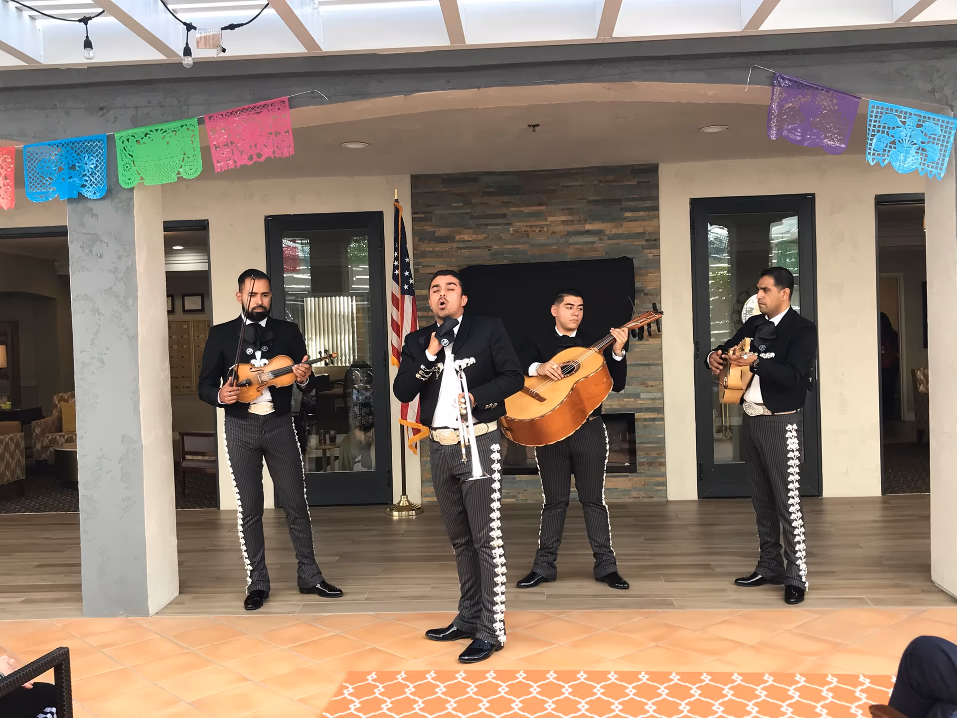 A mariachi band of four men dressed in traditional black charro suits with silver decorations performing indoors in a common area with a tiled floor, stone accent wall, and American flag in the background. Colorful papel picado decorations hang from the ceiling.