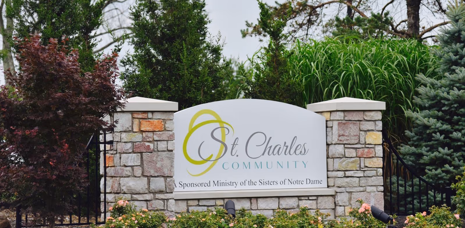 Stone sign for St. Charles Community surrounded by greenery and flowers, with trees and shrubs in the background.