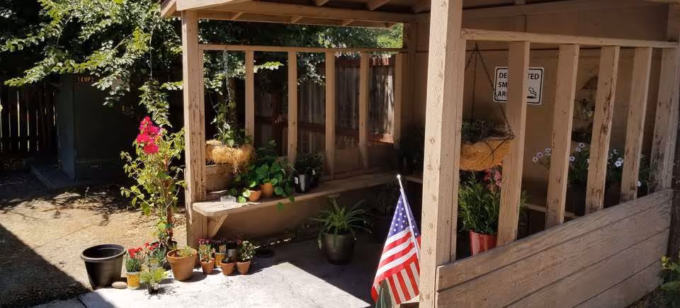 Outdoor covered patio area with wooden beams and a variety of potted plants, including flowers and greenery. An American flag is placed near the plants. A 'Designated Smoking Area' sign is visible on the wall in the background. Sunlight casts shadows on the concrete floor and surrounding area.