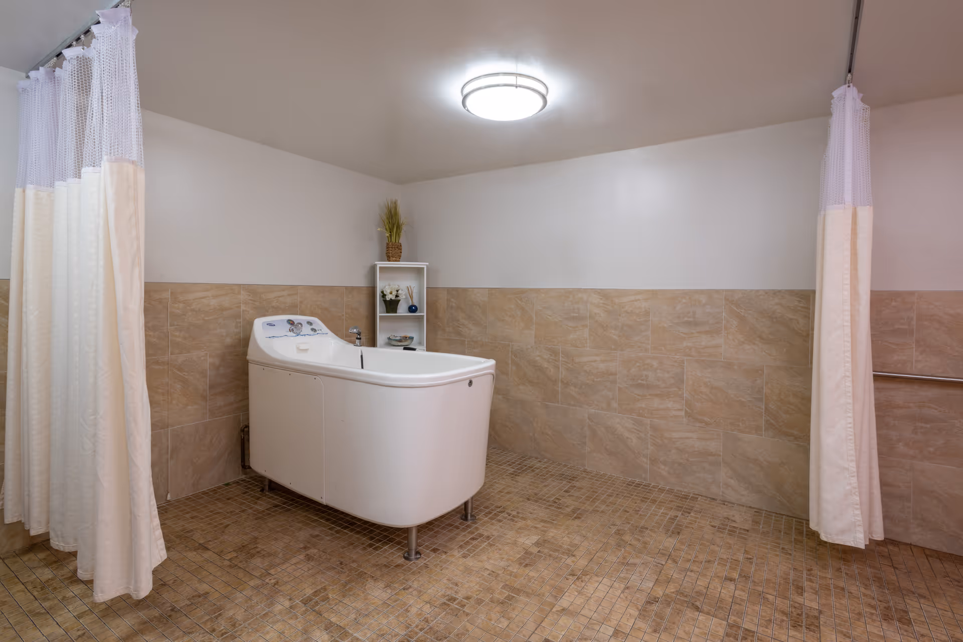 A spacious bathroom with beige tiled walls and floor, featuring a white walk-in bathtub in the center. There are two white privacy curtains on either side of the bathtub and a small white shelving unit with decorative items in the corner. The ceiling has a round light fixture.