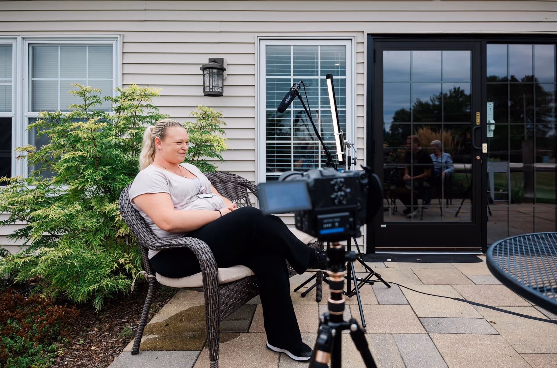A woman sits on a wicker chair on a paved patio outside a building while a camera and microphone are set up in front of her.