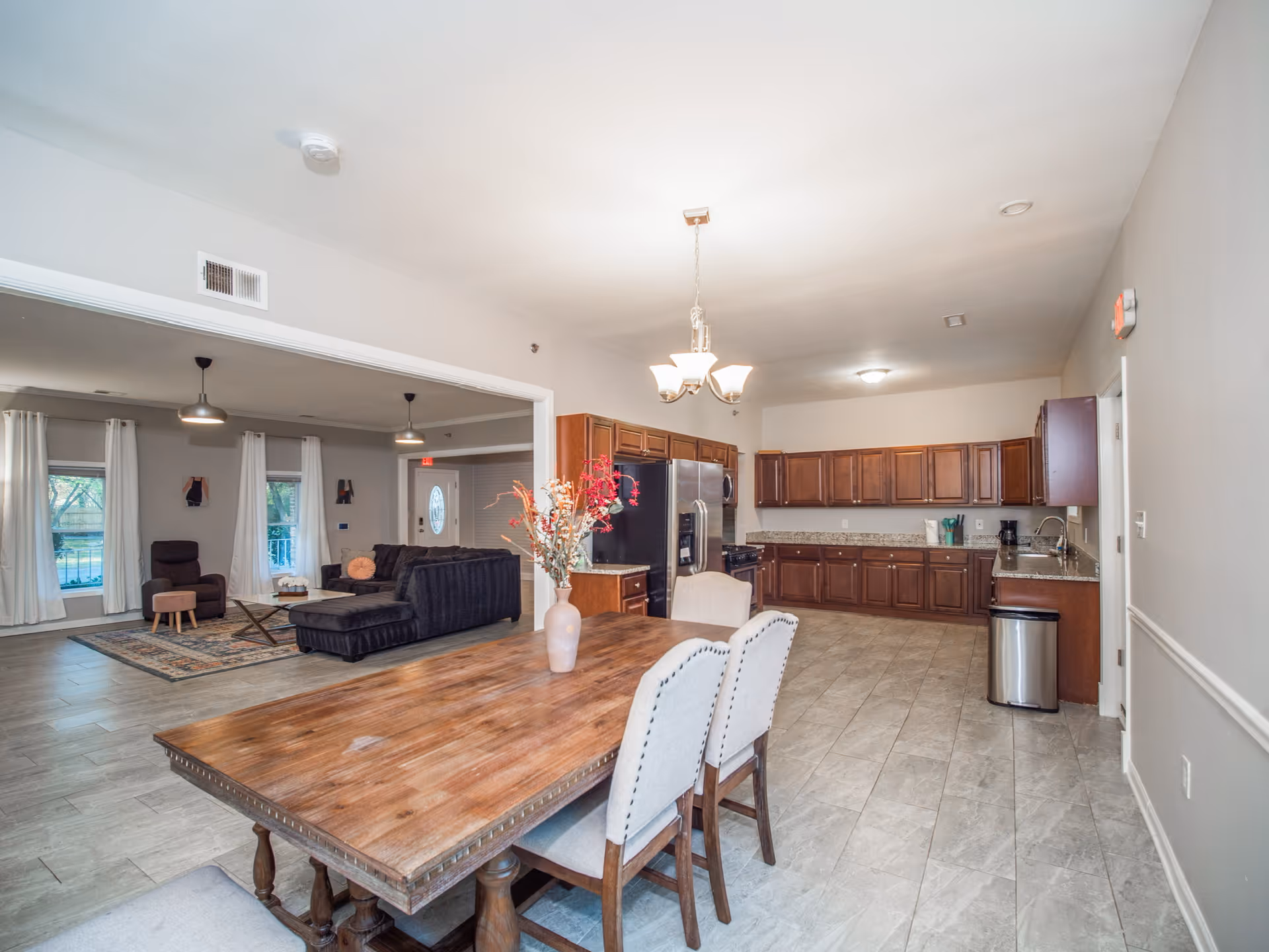 A spacious open-plan interior featuring a dining area with a wooden table and four upholstered chairs, a kitchen with wooden cabinets, granite countertops, stainless steel appliances, and a living room with a black sectional sofa, armchair, and windows with white curtains.