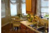 A cozy kitchen and dining area with wooden cabinets, a double sink, and a small round dining table with four chairs. The dining table is set with plates and a centerpiece of flowers. The room has windows with white curtains allowing natural light to brighten the space.