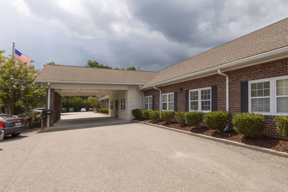 Exterior view of a brick building with white-framed windows and black shutters, a covered entrance area, neatly trimmed bushes along the building, a parked car on the left, and an American flag on a flagpole. The sky is cloudy and overcast.